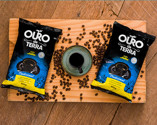 Dois pacotes de Café Ouro da Terra Tradicional numa tábua de madeira, com grãos de café e uma xícara no centro
