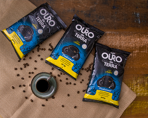 Três pacotes de Café Ouro da Terra sobre saco de juta, com xícara de café e grãos espalhados